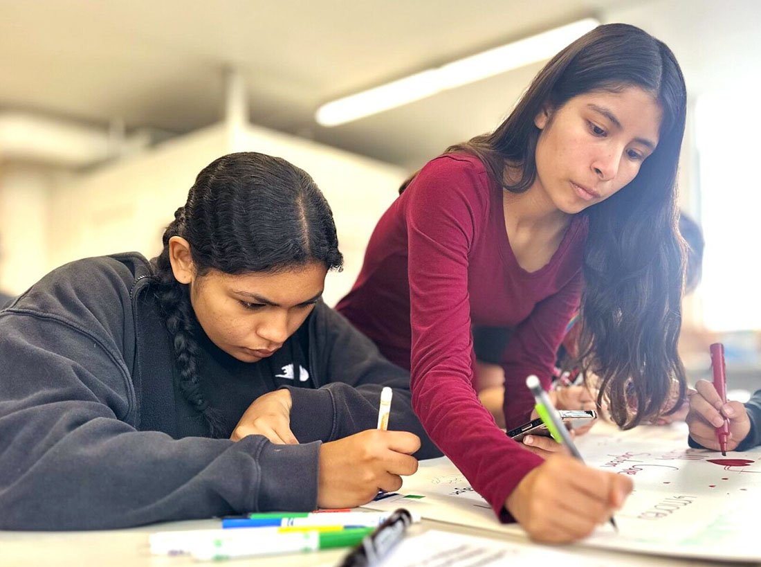 Two youth lean over a table, drawing with pens